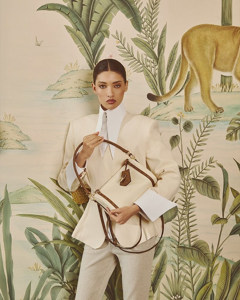 Model Aqua Parios holding a cream handbag wearing a cream blazer against a tropical painted wall mural