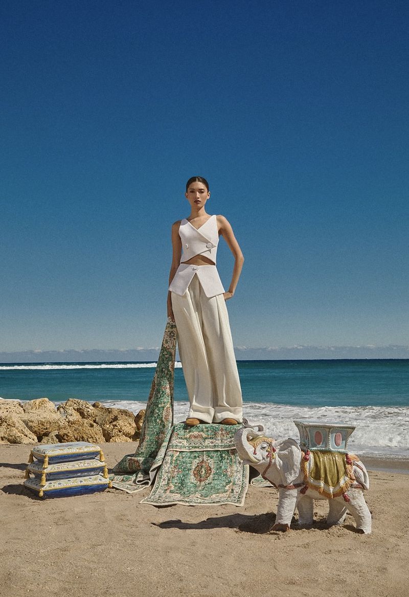 Model wearing a cream two-piece vest and trousers standing on a patterned rug on a sandy beach under a clear blue sky