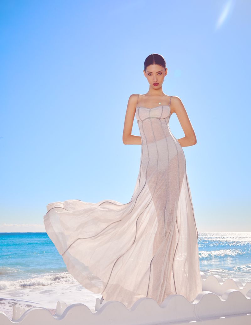 Model Aqua Parios wearing a long white dress standing against a bright blue ocean backdrop for Haute Living editorial