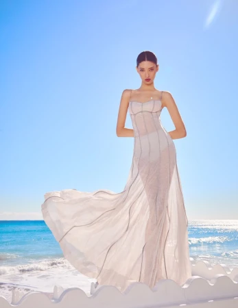 Model Aqua Parios wearing a long white dress standing against a bright blue ocean backdrop for Haute Living editorial