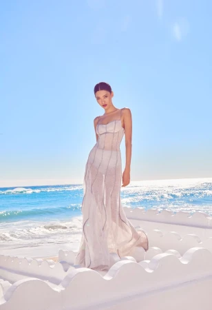 Model wearing a long sheer white dress standing on a white architectural terrace against an ocean and sky background.