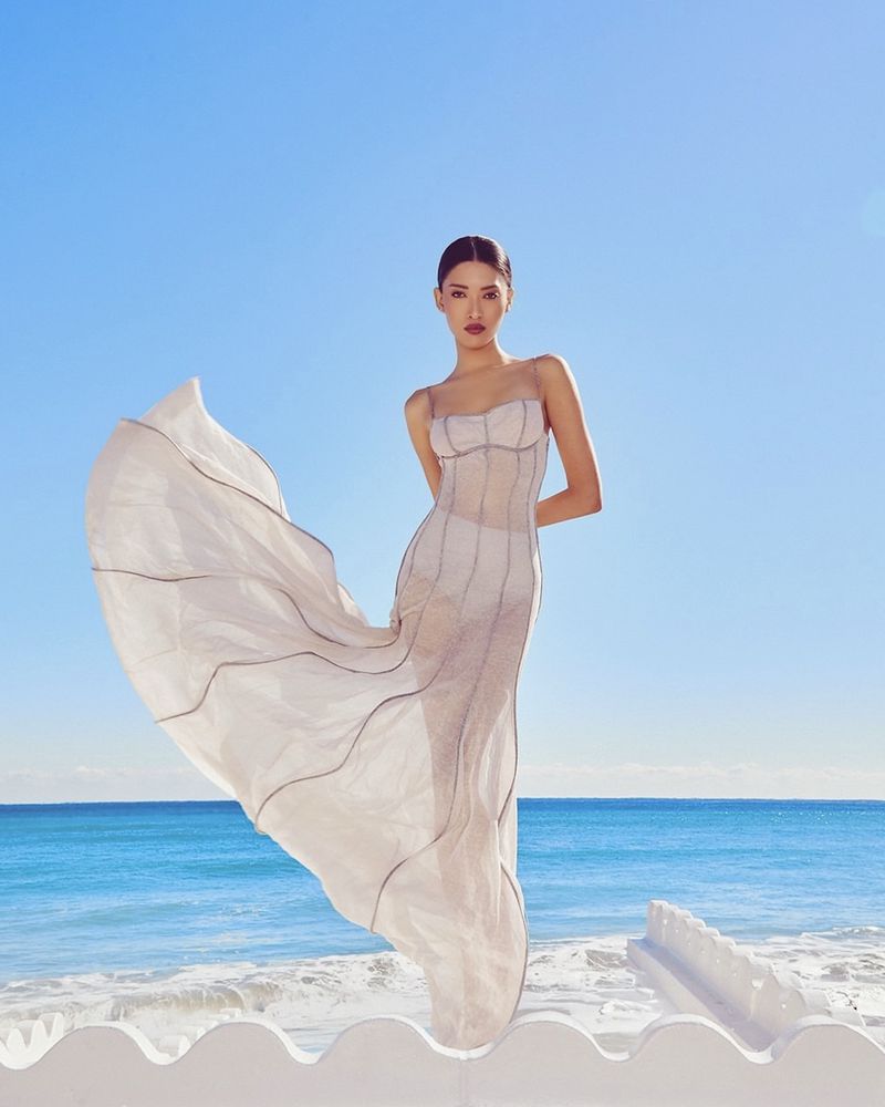 Model wearing a structured white dress standing against a bright blue ocean and sky background.