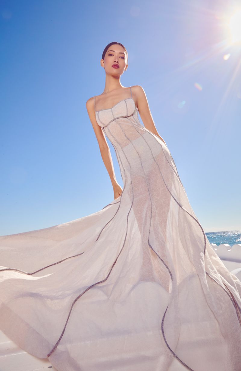 Model wearing a flowing white gown standing against a bright blue sky and ocean backdrop with sunlight flare.