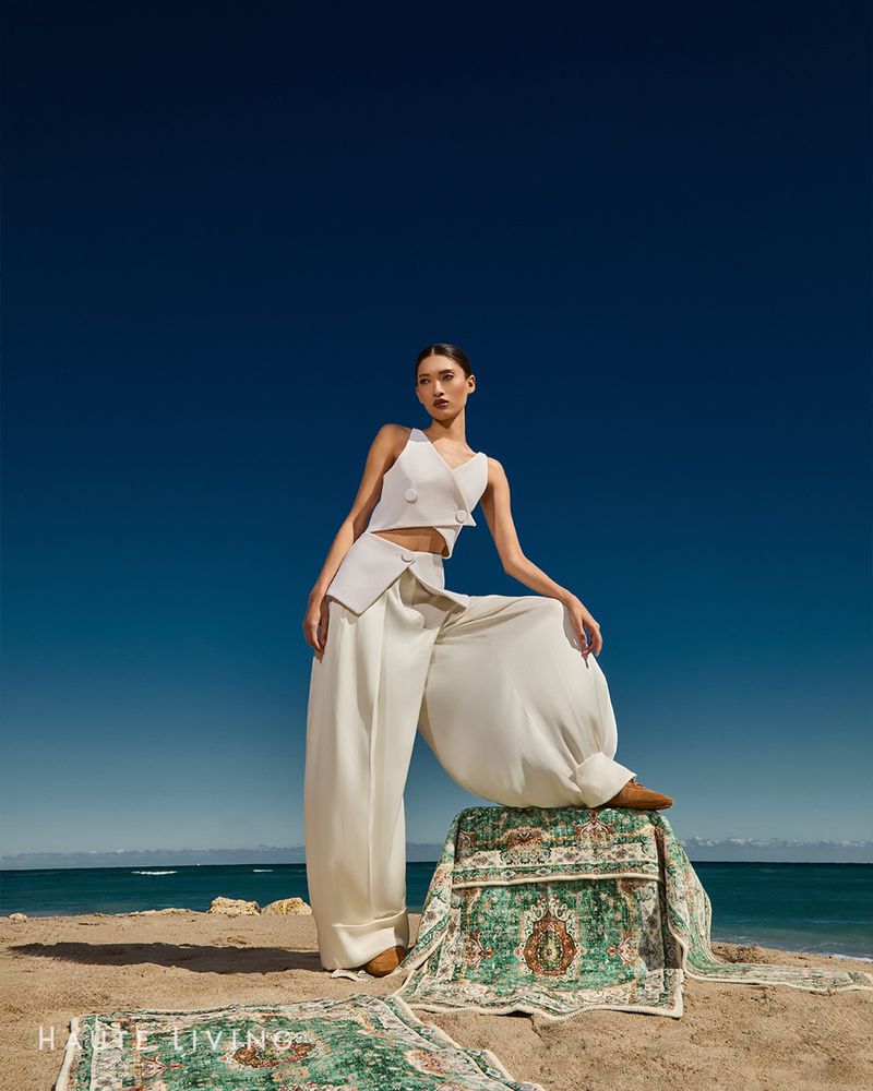 Model wearing white sleeveless top and wide trousers resting foot on patterned rug on sandy beach against clear sky