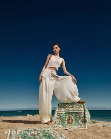 Model wearing white sleeveless top and wide trousers resting foot on patterned rug on sandy beach against clear sky