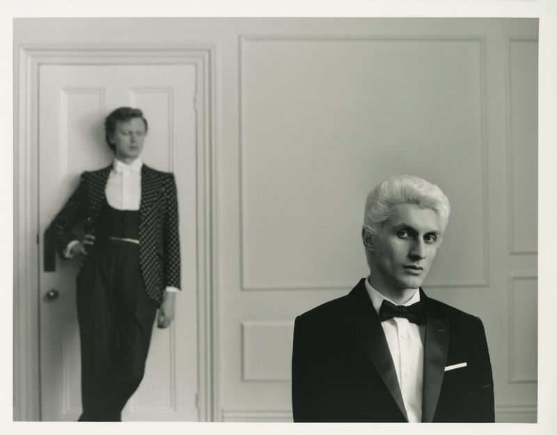 Two men in formal wear pose indoors against a white paneled wall. One man wears a dotted blazer, the other a classic tuxedo.