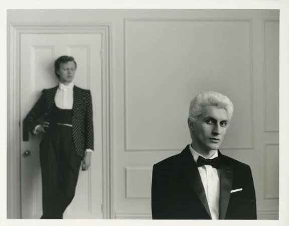 Two men in formal wear pose indoors against a white paneled wall. One man wears a dotted blazer, the other a classic tuxedo.
