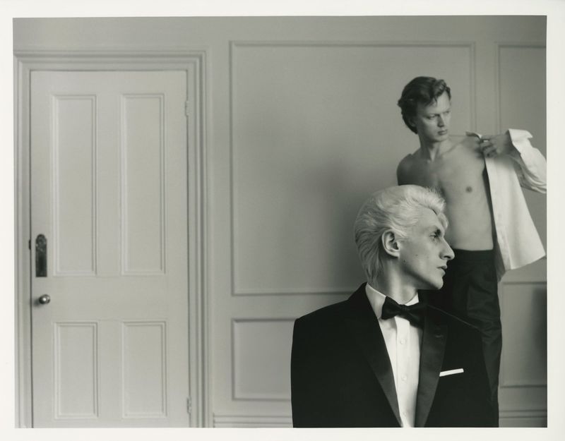 Black and white studio portrait of two adult males, one in a tuxedo, the other removing his shirt in front of a light-colored wall.