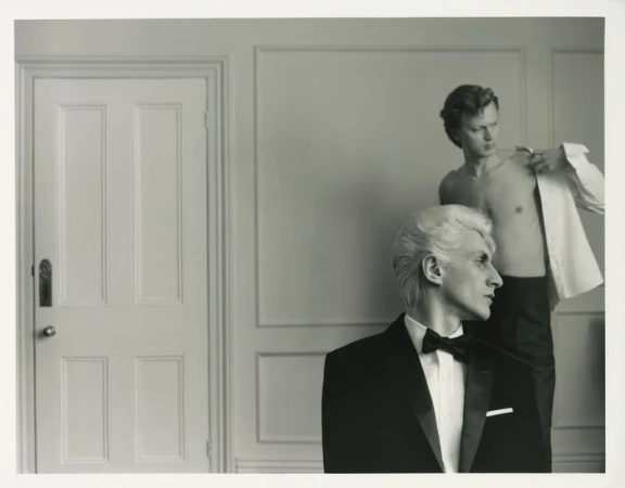 Black and white studio portrait of two adult males, one in a tuxedo, the other removing his shirt in front of a light-colored wall.