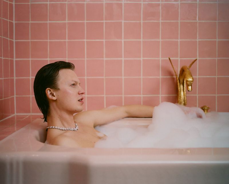A man wearing a diamond necklace relaxes in a bubble-filled bathtub against a pink tile wall with a gold faucet shaped like a swan.