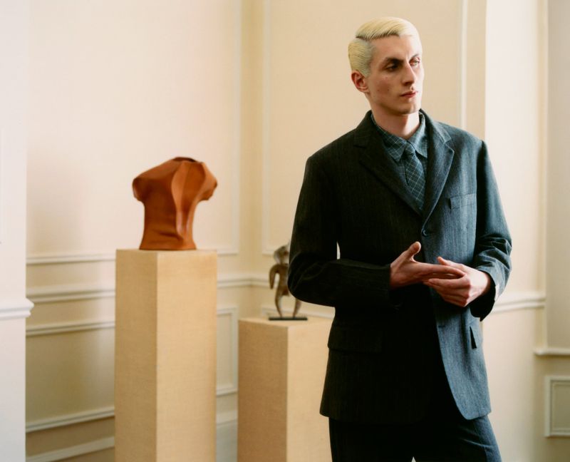 An adult male with a fair complexion poses in a grey suit jacket with sculptures on pedestals in a gallery-like interior setting.