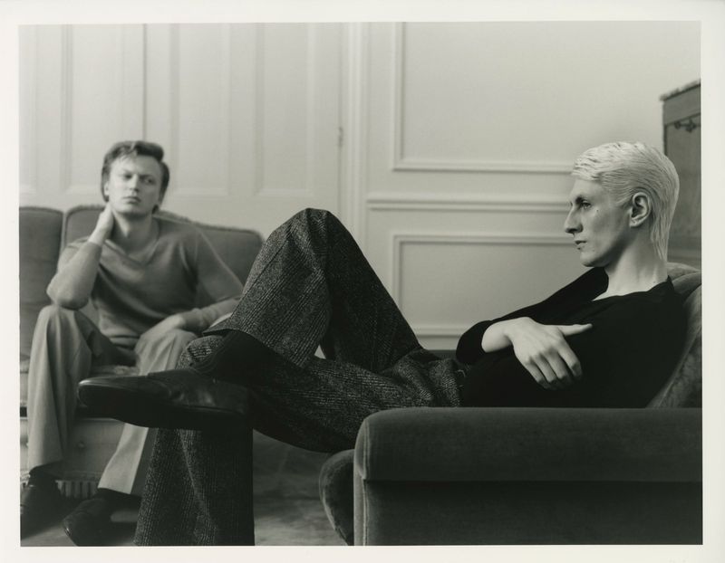 Two adult men in a monochromatic indoor shot, one seated on a sofa and the other in an armchair, looking away with a neutral expression.