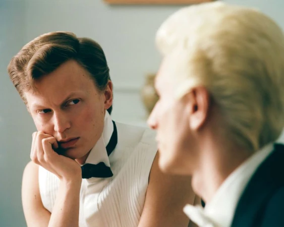 Two adult males in formal wear: one faces forward with a pensive expression, and the other is in partial view with blonde hair.