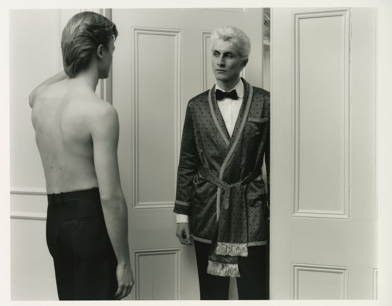 Two men indoors: one shirtless wearing trousers, the other dressed in a robe with a bow tie, standing in a doorway, black and white.