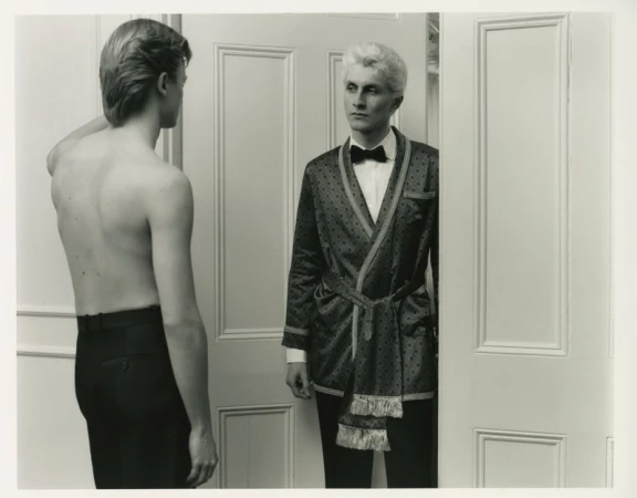 Two men indoors: one shirtless wearing trousers, the other dressed in a robe with a bow tie, standing in a doorway, black and white.