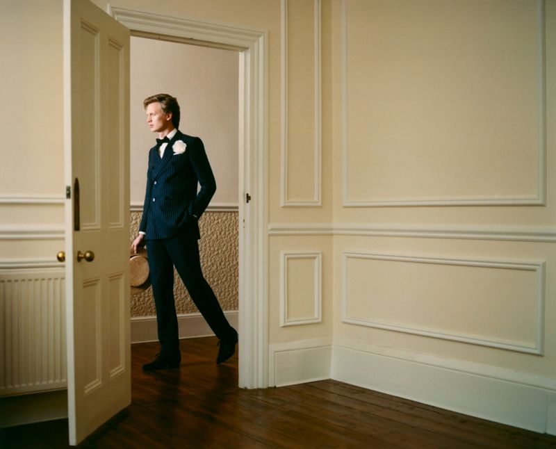 Stylish adult male in a pinstripe suit walks through a doorway holding a boater hat indoors with decorative molding on the walls.