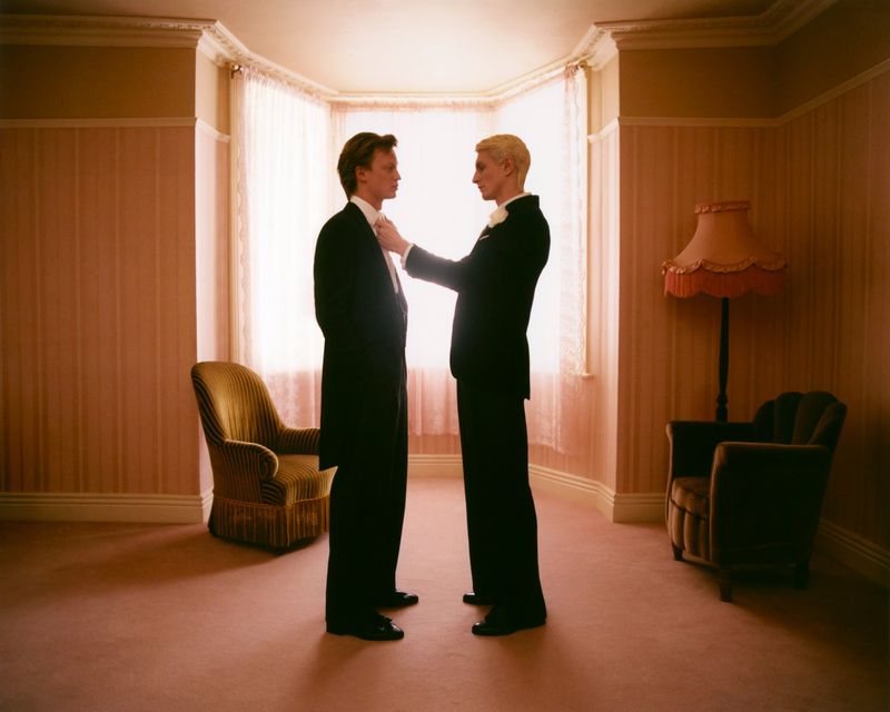 Two adult males in formal wear face each other in a room with pink patterned wallpaper and a bay window, adjusting each other's collars.