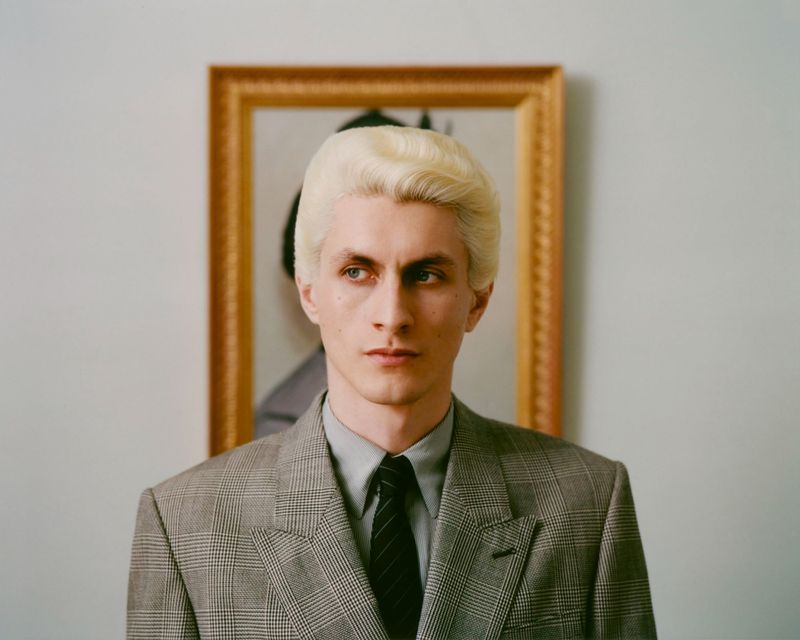 Headshot of adult male with bleached blond hair wearing grey plaid suit and tie, posed in front of portrait in gold frame.
