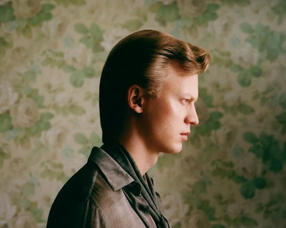 Portrait of a blond male with a pompadour hairstyle in a side profile view, wearing a collared shirt, with a floral wallpaper background.