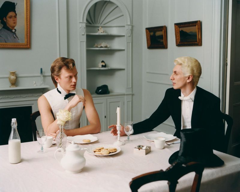 Two men in formalwear at a table with food, drinks, and a candle. The scene has paintings and a china cabinet.