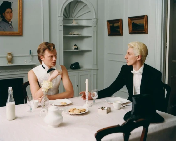 Two men in formalwear at a table with food, drinks, and a candle. The scene has paintings and a china cabinet.