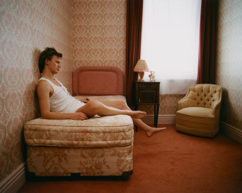 Man lounges on patterned bed in a room with floral wallpaper, table lamp, armchair, and red carpet, bathed in soft window light.