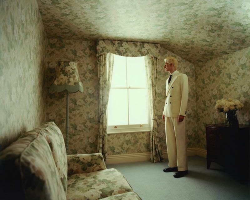 A man stands in a room with floral wallpaper, wearing a white suit and black shoes, beside a window and sofa, capturing a moment.