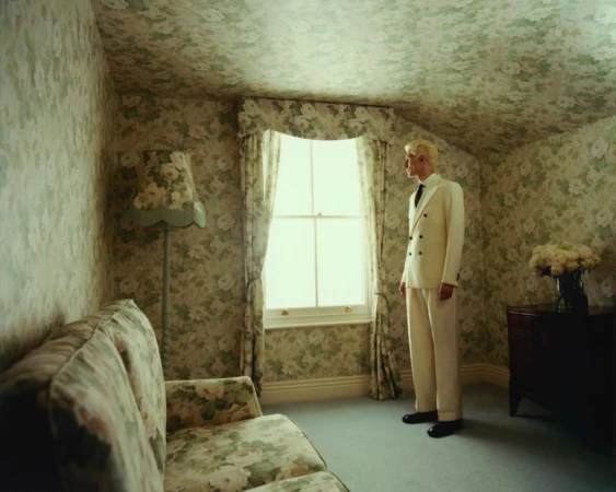 A man stands in a room with floral wallpaper, wearing a white suit and black shoes, beside a window and sofa, capturing a moment.