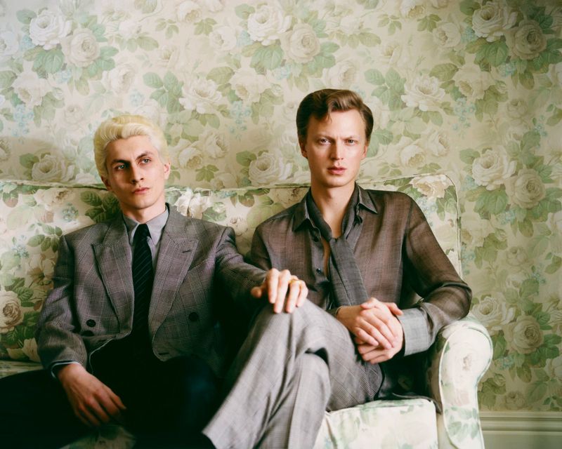 Two adult men pose on a floral couch indoors. One man with blond hair sits in a grey suit. A man with brown hair sits in a grey suit.