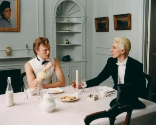 Jonas Glöer and Henry Kitcher in formal eveningwear seated at a dining table set with tea and biscuits.