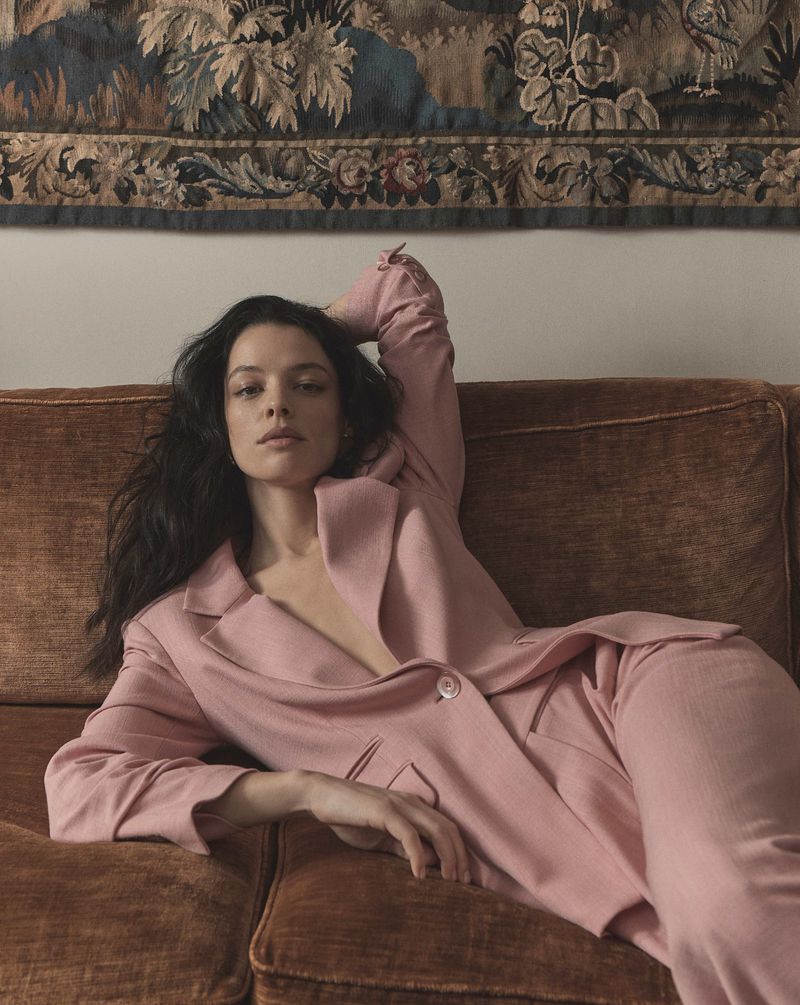 Model Nigina Sharipova wearing a pink pantsuit reclining on a brown sofa beneath a hanging tapestry
