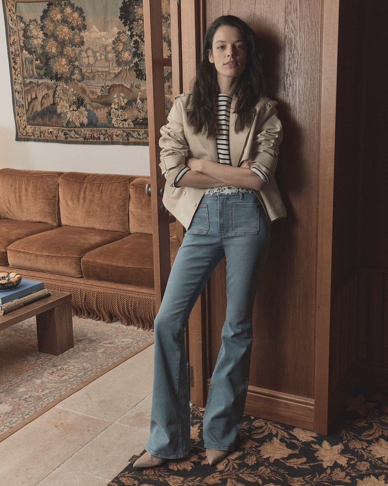 A woman stands indoors wearing a tan blazer, striped shirt, and light-wash denim bell-bottom jeans in a fashion editorial style shot.