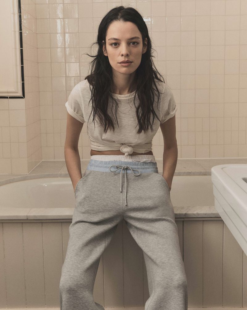 Model wearing a white t-shirt and grey sweatpants sitting in front of a tiled bathtub for Veronica Beard