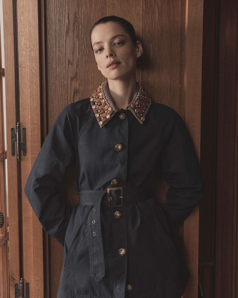 Model in a dark belted coat with an embellished collar posing against wood paneling for Veronica Beard Spring 2026