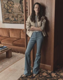 Model leaning against a wooden doorframe wearing a tan jacket, striped shirt, and flared blue jeans inside a furnished room.