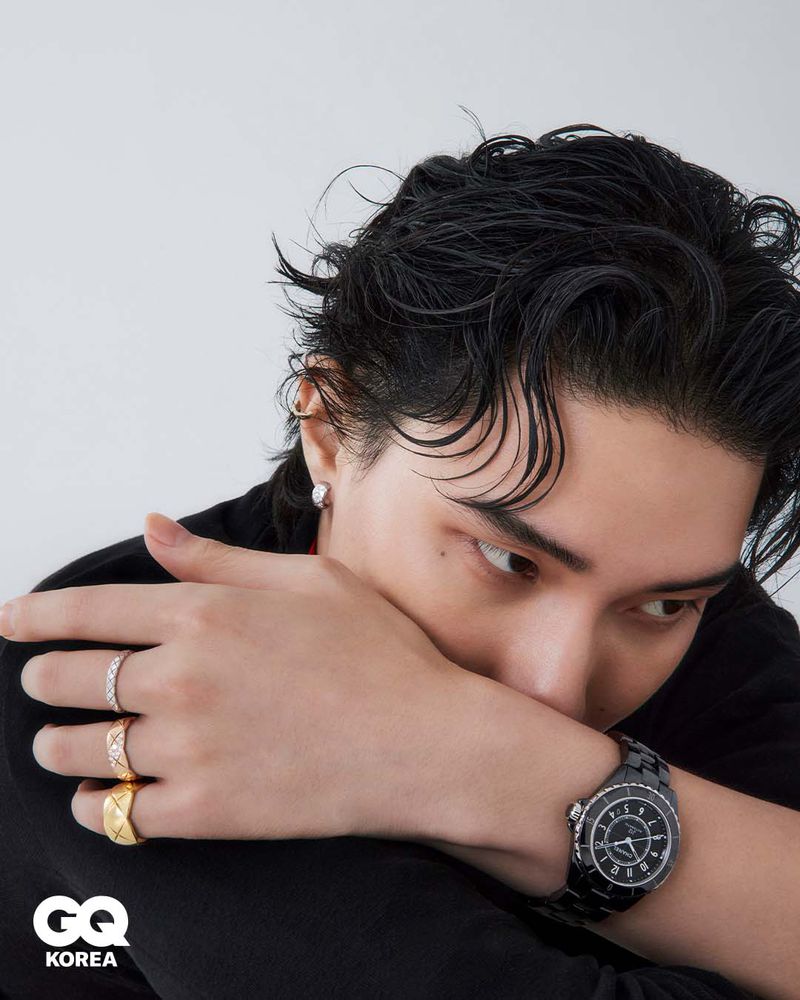 Close-up portrait of a model with wet dark hair wearing a black top, rings, and a dark wristwatch against a white background.