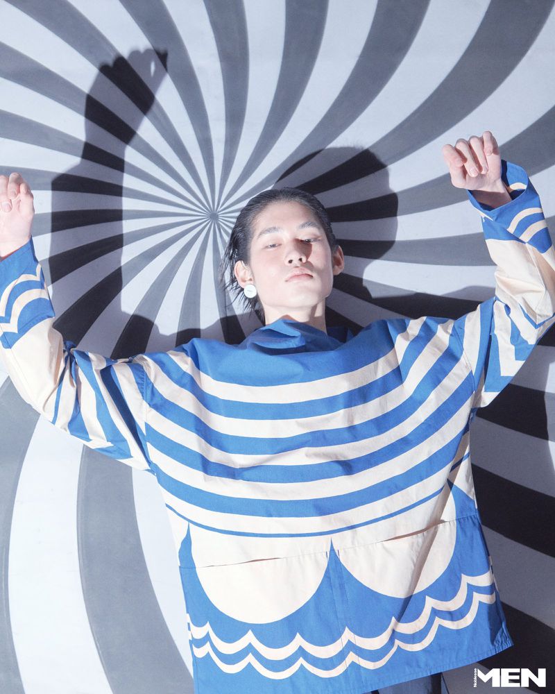 Model lying against a black and white striped spiral background wearing a blue and cream patterned top with arms raised