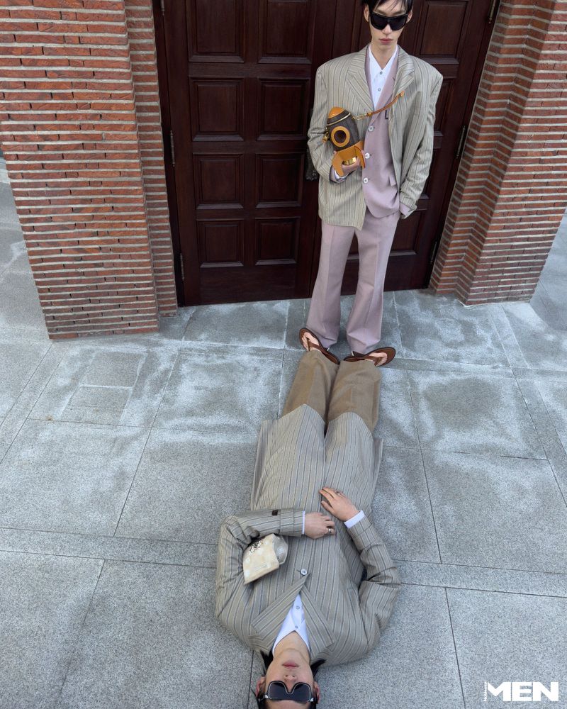 Two fashion models pose outdoors on a concrete surface near a brick wall and a dark wood door, wearing coordinated blazers and pants.