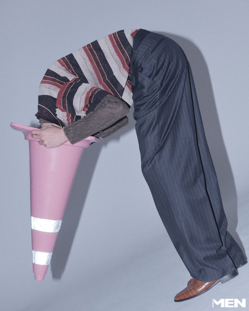 Model bent backward wearing a striped sweater and trousers with head inside a pink traffic cone against grey background