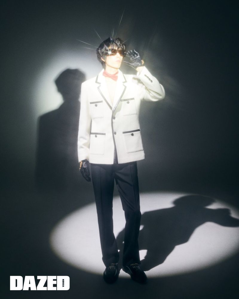 A full-length studio portrait of an adult male in an ivory jacket and dark pants, wearing sunglasses, with light shining on him.