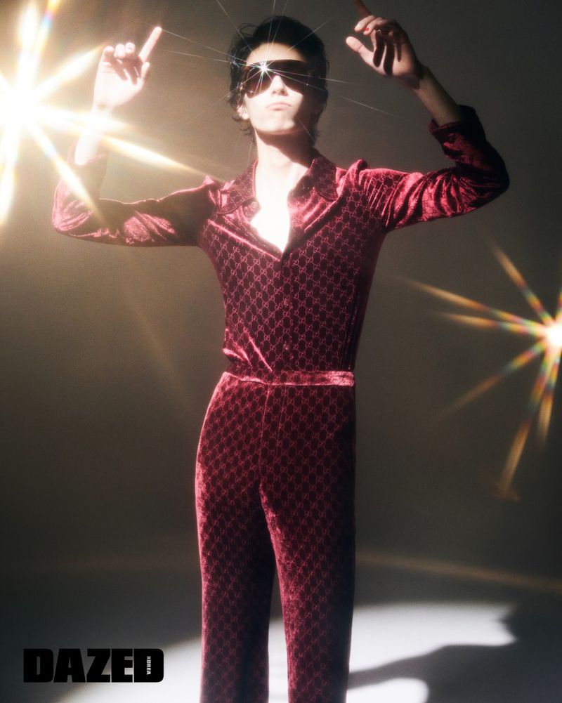 Model wearing a patterned burgundy velvet jumpsuit and oversized sunglasses posing with arms raised under starburst lens flares