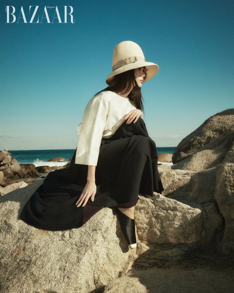 Model wearing a wide-brimmed hat, white top, and dark skirt seated on coastal rocks against an ocean and clear blue sky backdrop