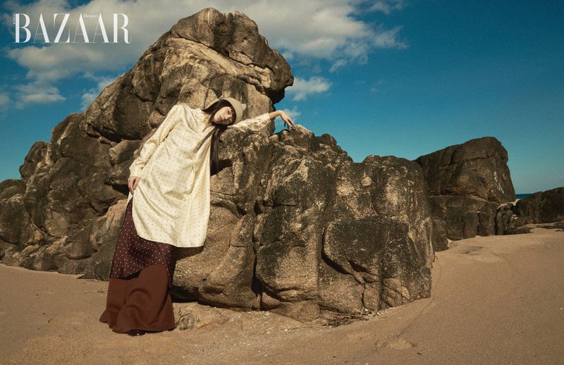 Model leaning against a large rock formation on a sandy beach under a clear blue sky for Harper's Bazaar Korea editorial