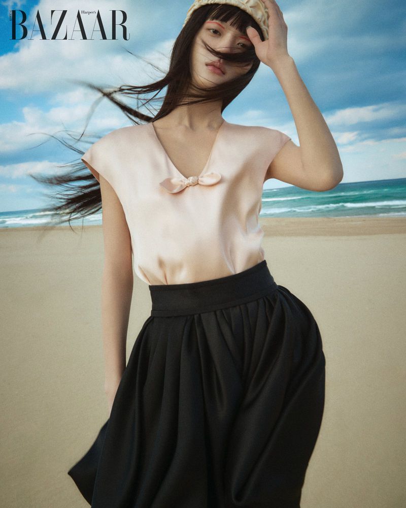 Model in a pale pink top and black skirt posing on a sandy beach with the ocean and a cloudy sky in the background.