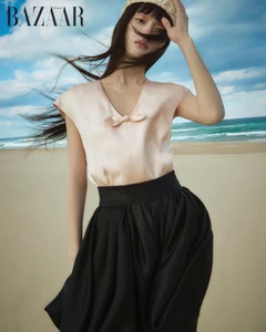 Model in a pale pink top and black skirt posing on a sandy beach with the ocean and a cloudy sky in the background.