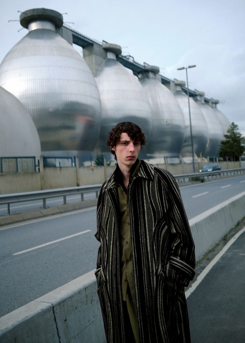 Male model wearing a striped coat standing on a roadside with industrial spherical structures in the background.