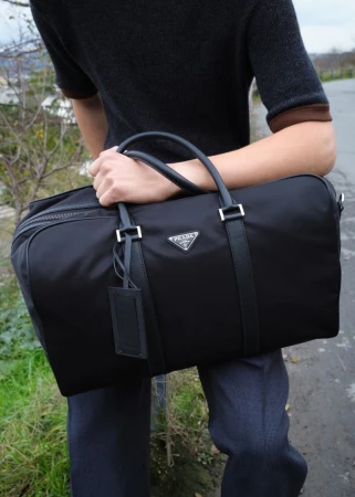 Man holding a black duffel bag wearing a dark short-sleeve top on an outdoor path