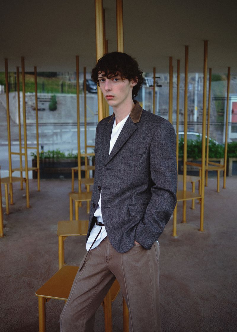 A young male model posing outdoors among thin golden architectural poles wearing a grey tweed jacket and brown trousers.