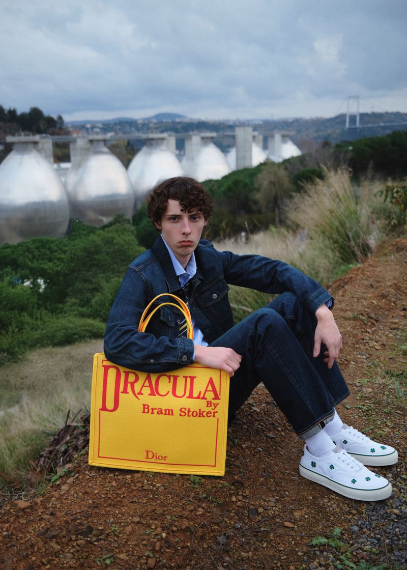 A young male model seated on a hillside outdoors wearing a denim jacket and holding a large yellow tote bag that reads Dracula.