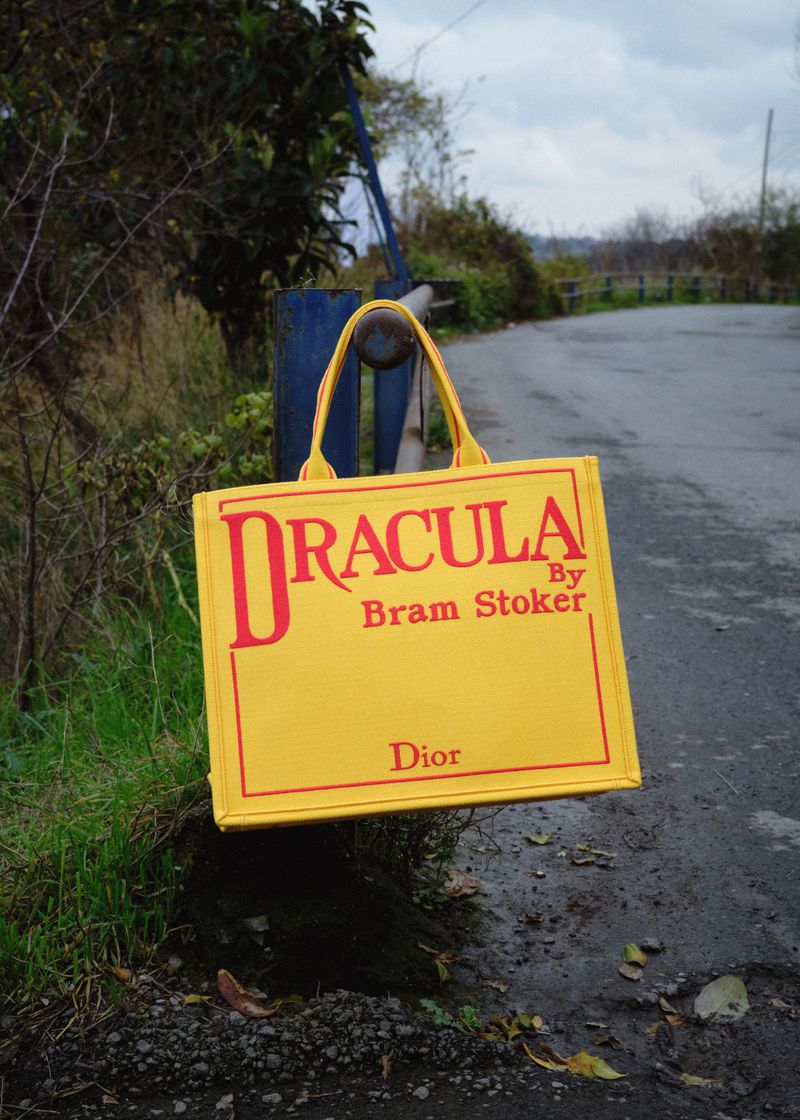 Bright yellow canvas tote bag with red Dracula text hanging on a roadside metal rail in an overcast outdoor setting